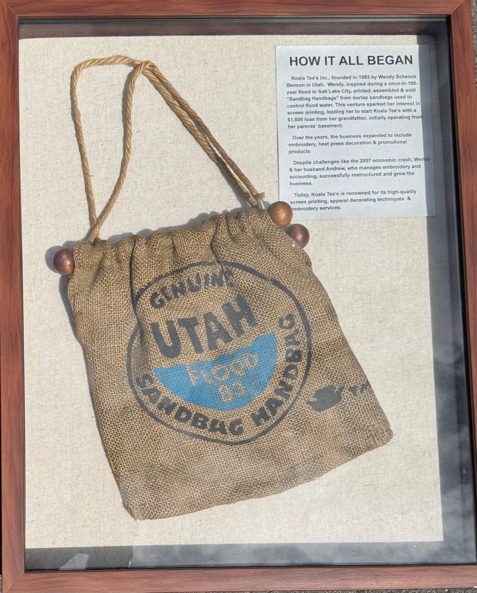 The original 'Genuine Utah Sandbag Handbag - Flood '83,' a burlap bag Koala-Tees printed and sold during the 1983 Salt Lake flood, framed in a shadow box beside a 'How It All Began' placard.