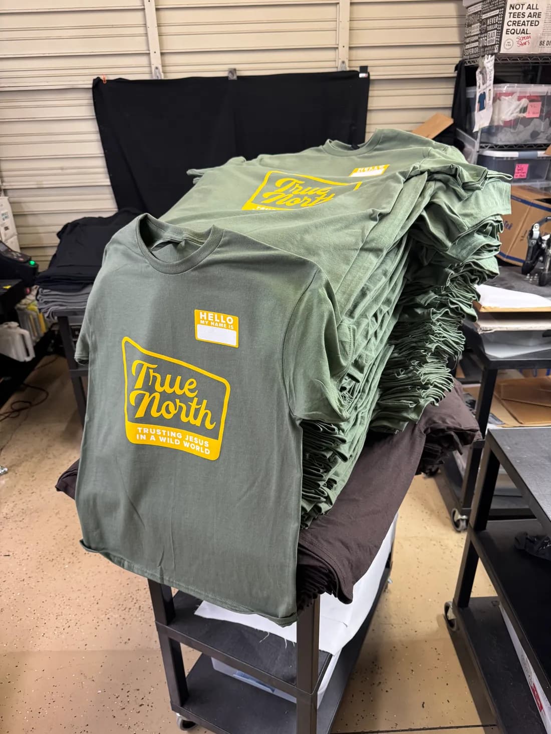 A tall stack of sage green Gildan tees with a two-color True North print for a local church group