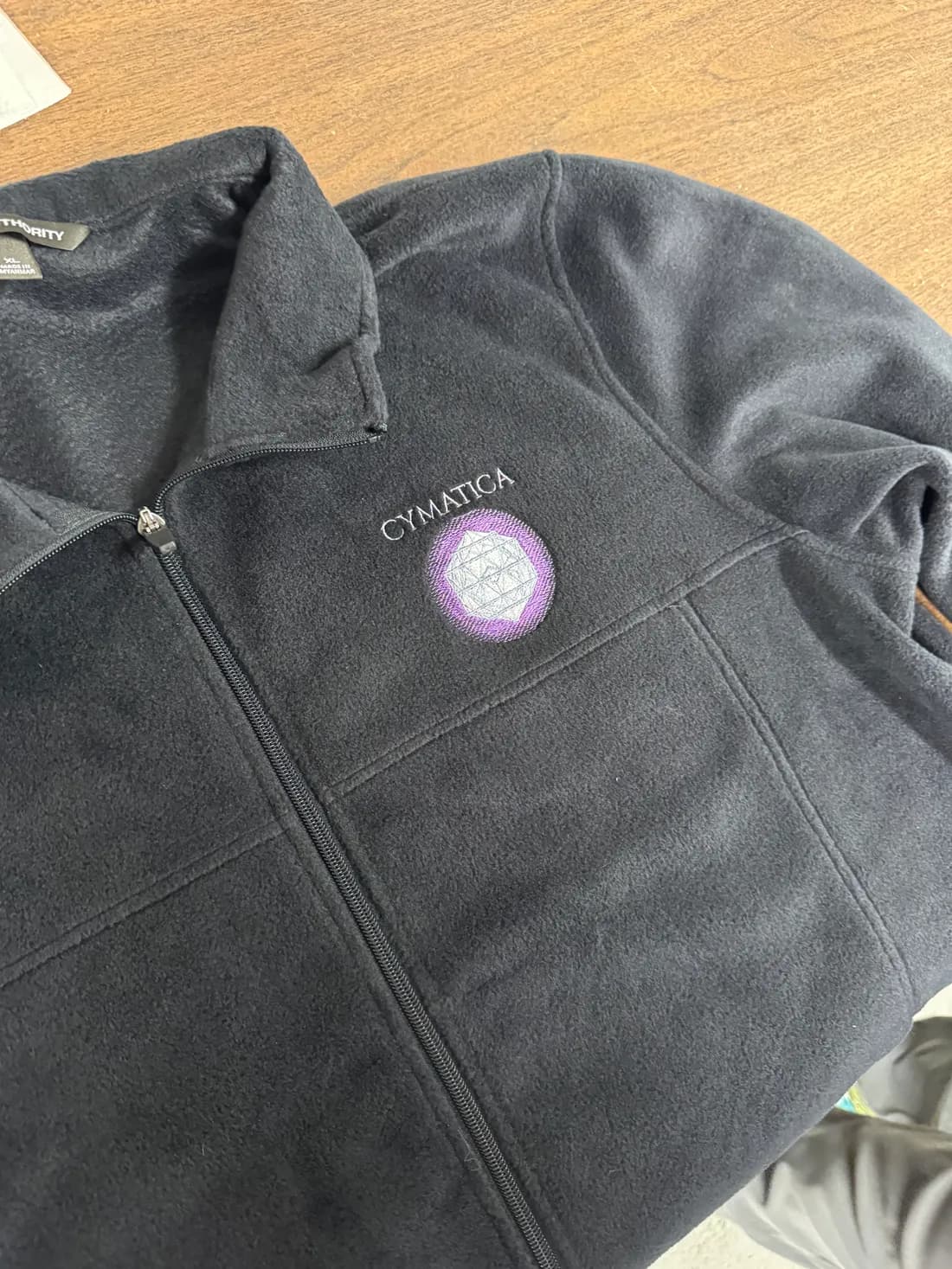 Dark full-zip microfleece with an embroidered Cymatica logo on the chest