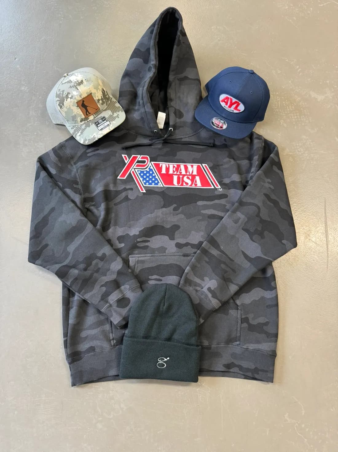 Black camo hoodie with an applique-embroidered XP Team USA logo, shown with custom caps and a beanie