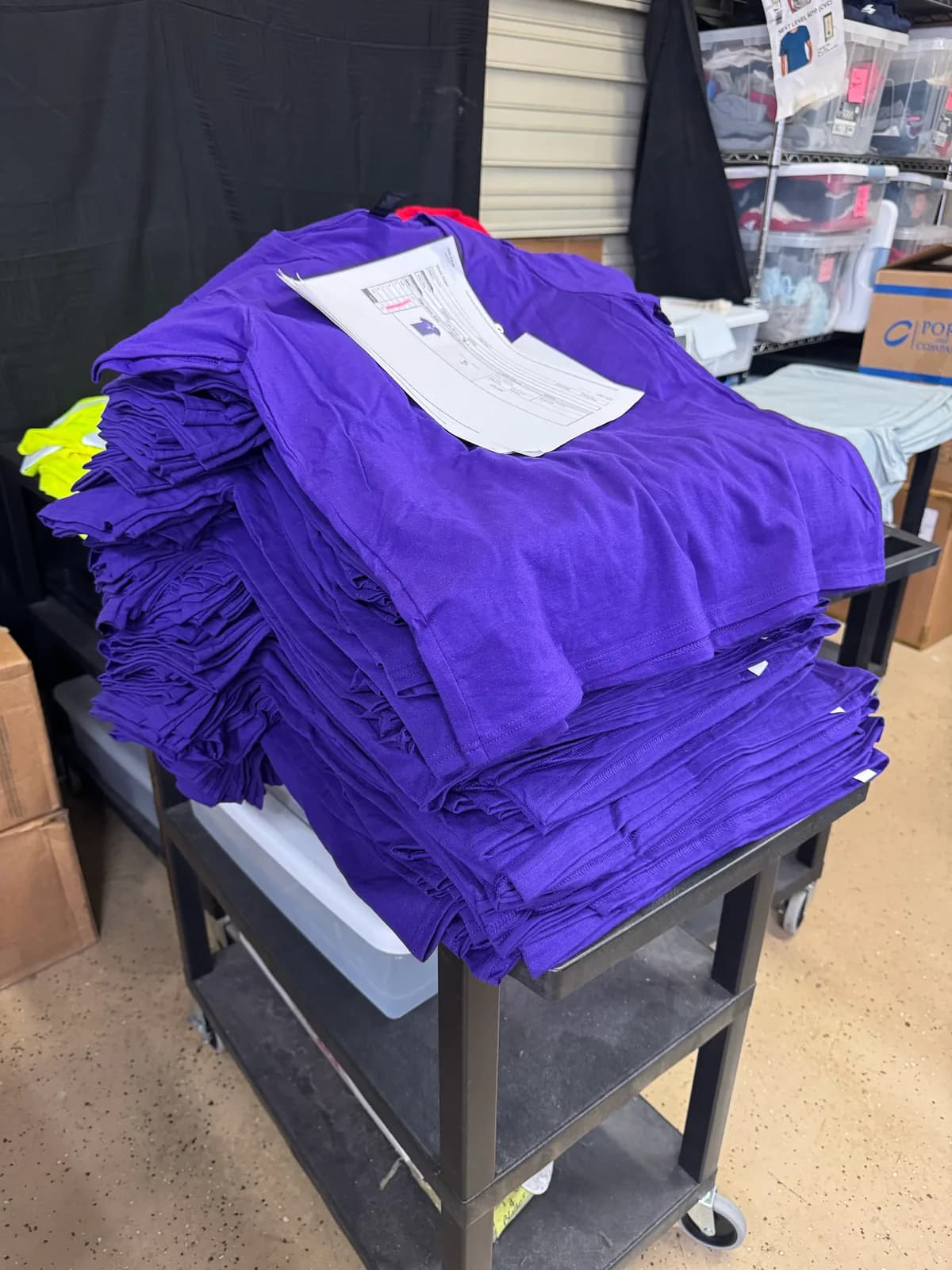 A tall stack of purple staff t-shirts staged on a shop cart with the work order on top, ready for printing