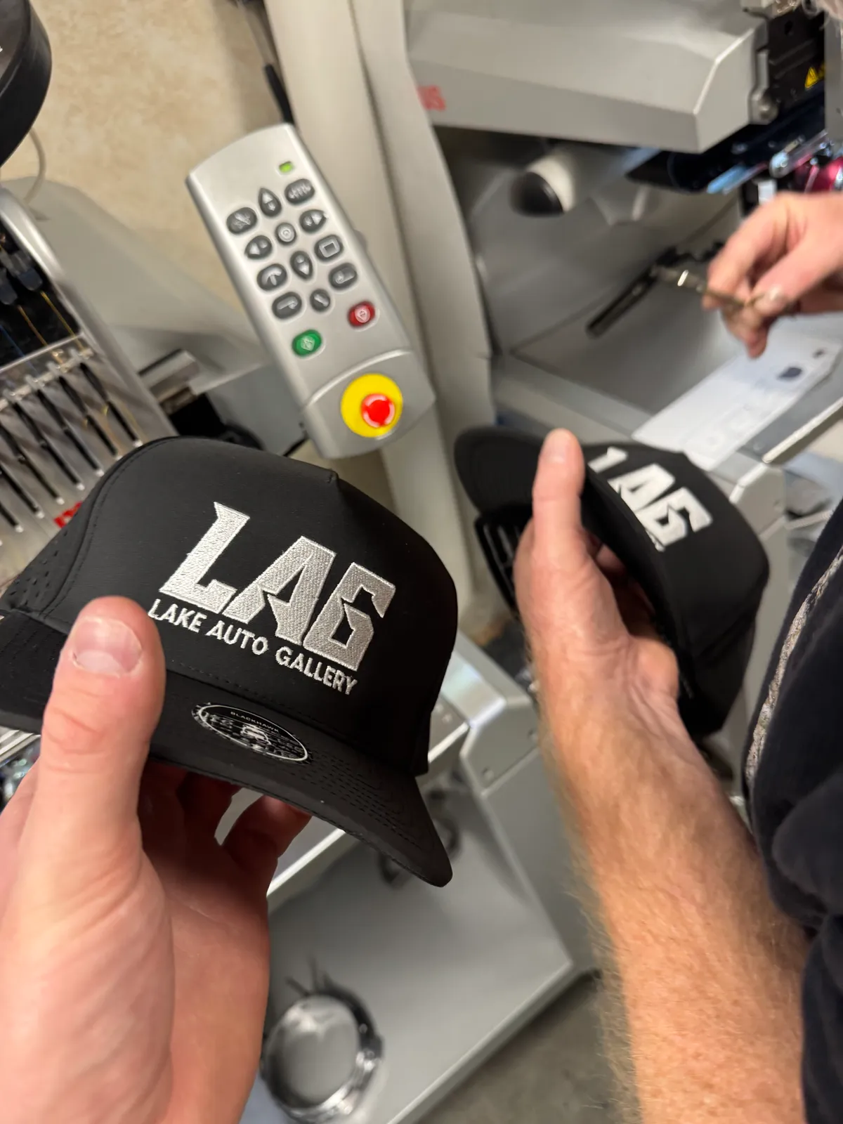 Two black caps with white embroidered Lake Auto Gallery logos, fresh off the embroidery machine