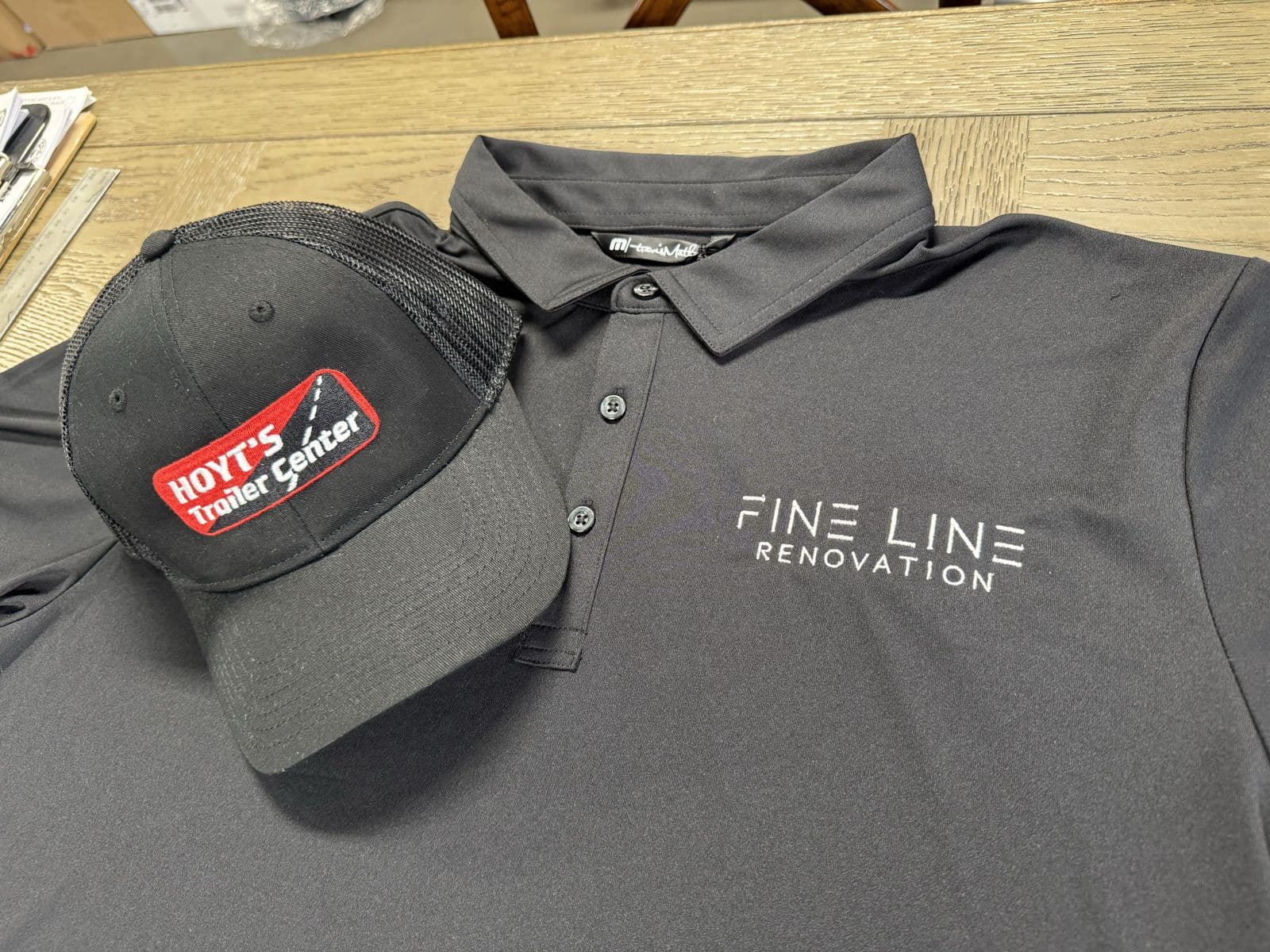 Embroidered Fine Line Renovation polo on the shop table with a Hoyt's Trailer Center patch trucker hat resting on it